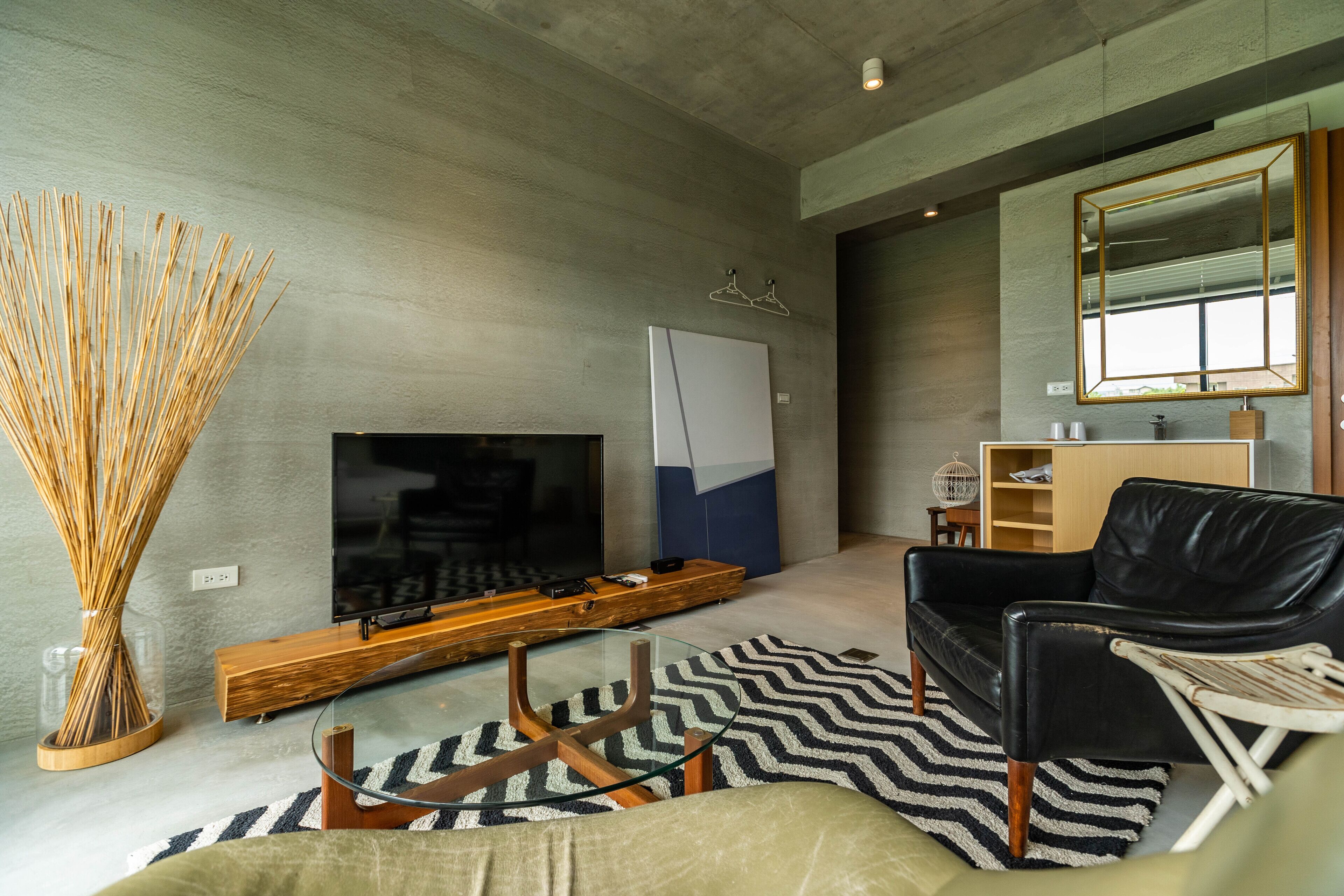 A modern living room with a flat-screen TV, a wooden entertainment unit, a black leather sofa, and a large mirror.
