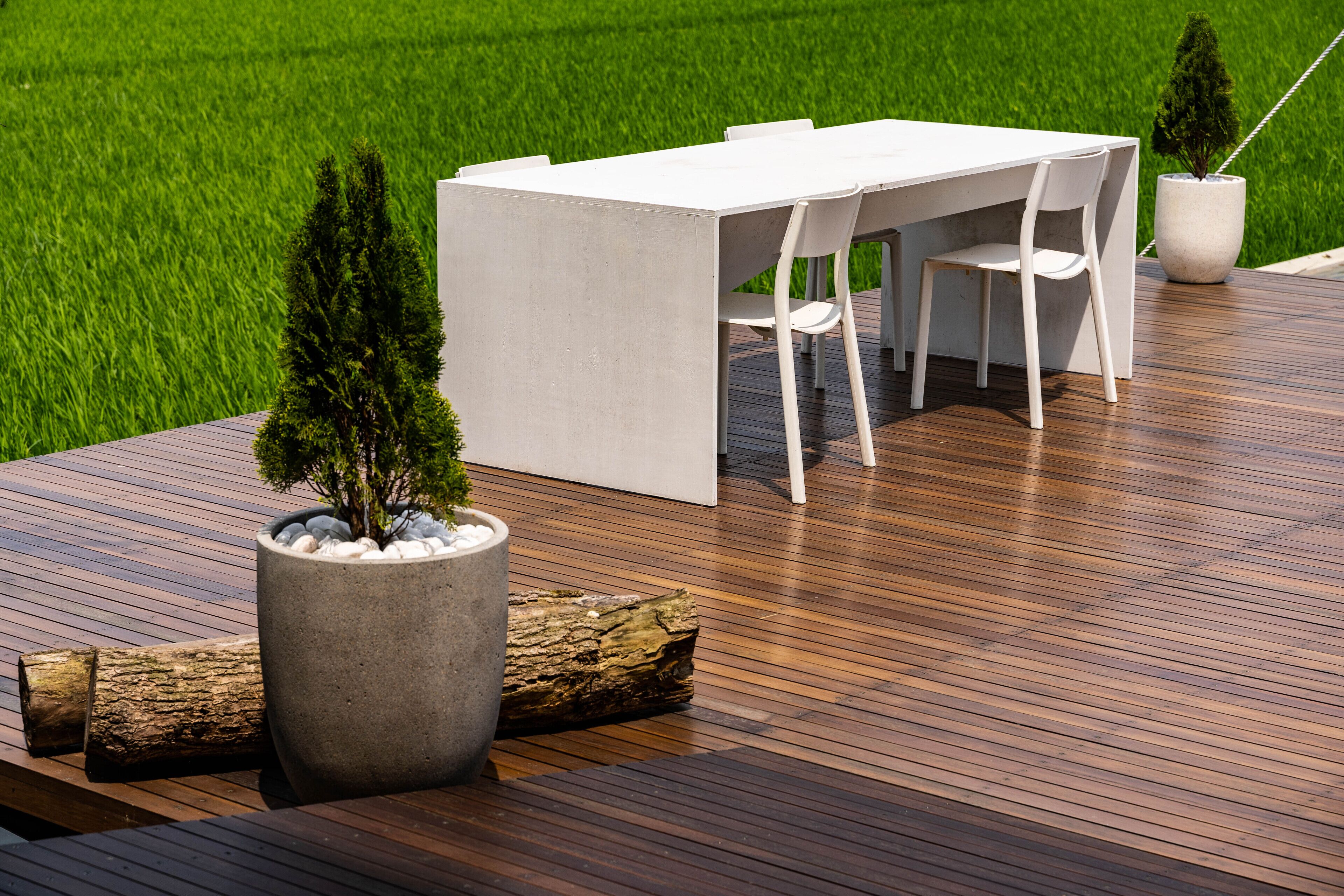 Un espace repas extérieur moderne, avec une table et des chaises blanches, entouré d’une terrasse en bois et de plantes en pot.