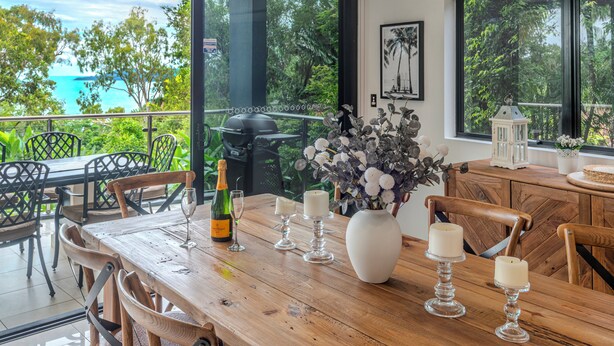 Une table à manger en bois, avec des chaises, un vase rempli de fleurs, des bougies et une bouteille de champagne.