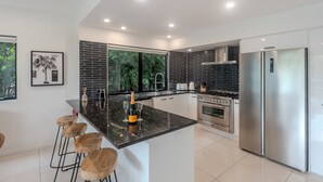 Une cuisine moderne dotée d’un îlot central, de tabourets de bar, d’un réfrigérateur en acier inoxydable et d’une grande fenêtre offrant une vue sur les arbres.