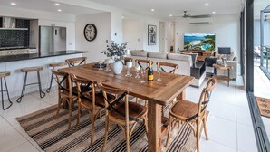 Une cuisine moderne équipée d’appareils électroménagers en acier inoxydable, un coin repas avec une table et des chaises en bois, et un salon doté d’une grande télévision et offrant une vue sur l’océan.