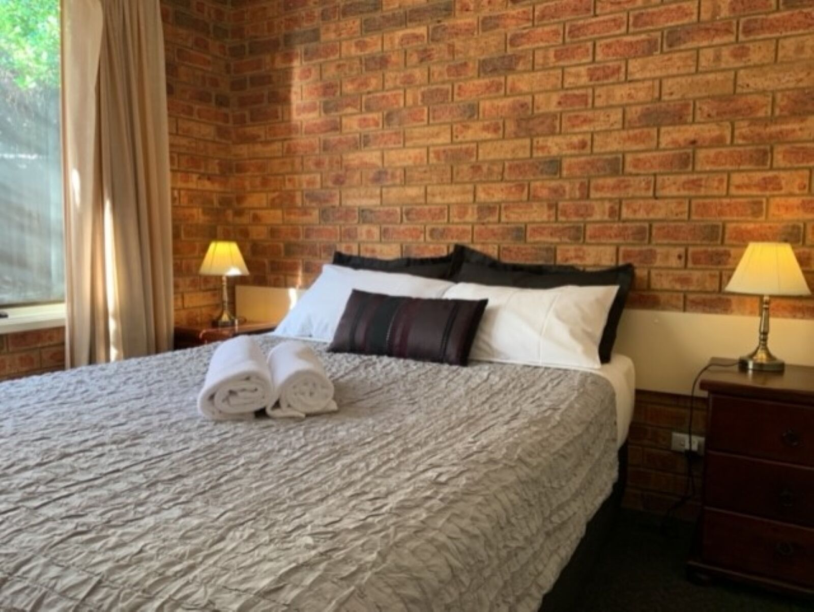 A hotel room with a brick wall, a bed with a grey quilt, two rolled towels, two bedside lamps, and a window with curtains.