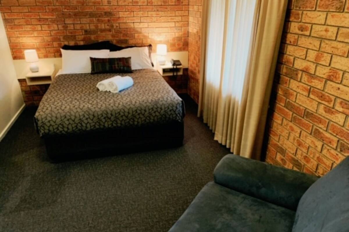 Una habitación de hotel con una pared de ladrillo, una cama con colcha estampada, una mesita de noche con lámpara y un sofá gris.