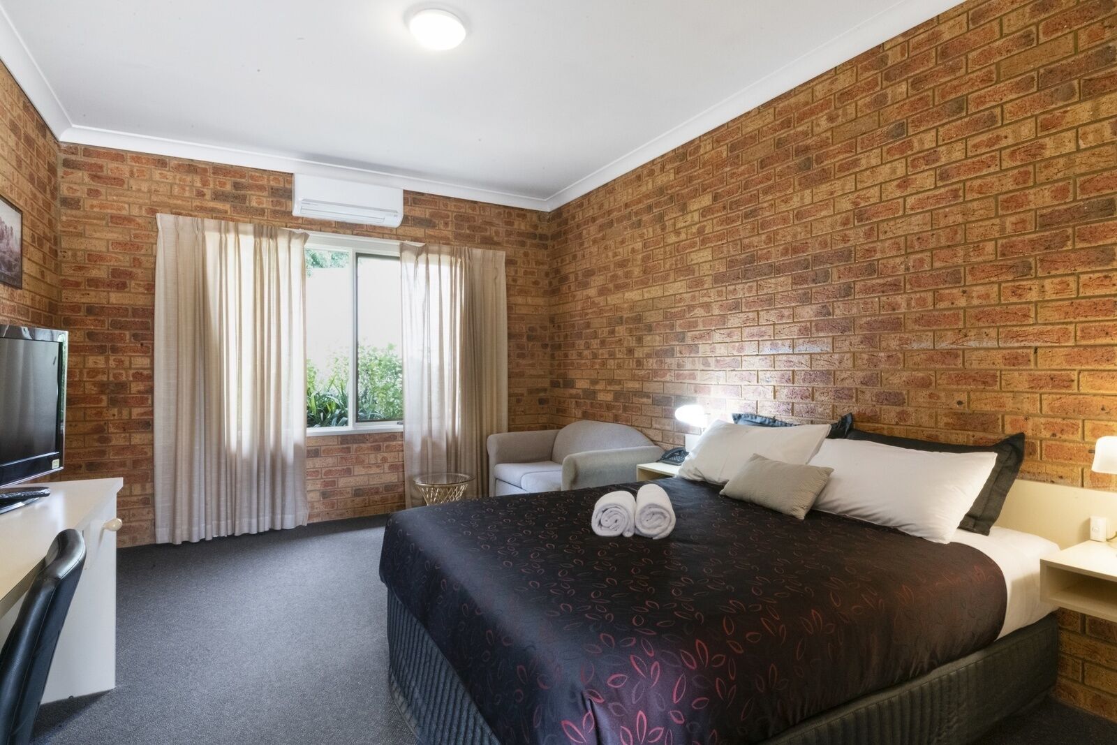 Une chambre d’hôtel avec un mur en briques, un lit recouvert d’une courtepointe à motifs sombres, un bureau avec une télévision et une fenêtre avec des rideaux.