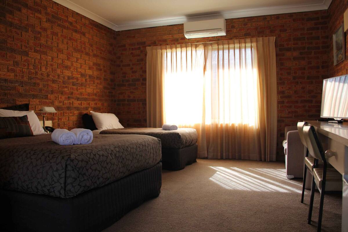 Habitación de hotel con pared de ladrillo, dos camas, un escritorio y un amplio ventanal con cortinas.