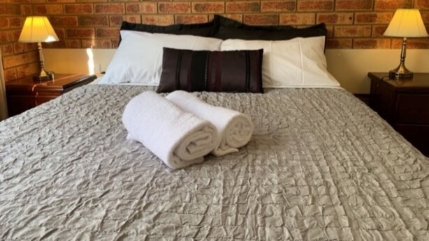 Una cama bien tendida con una toalla blanca enrollada, un almohadón blanco y negro, y una colcha gris con textura.