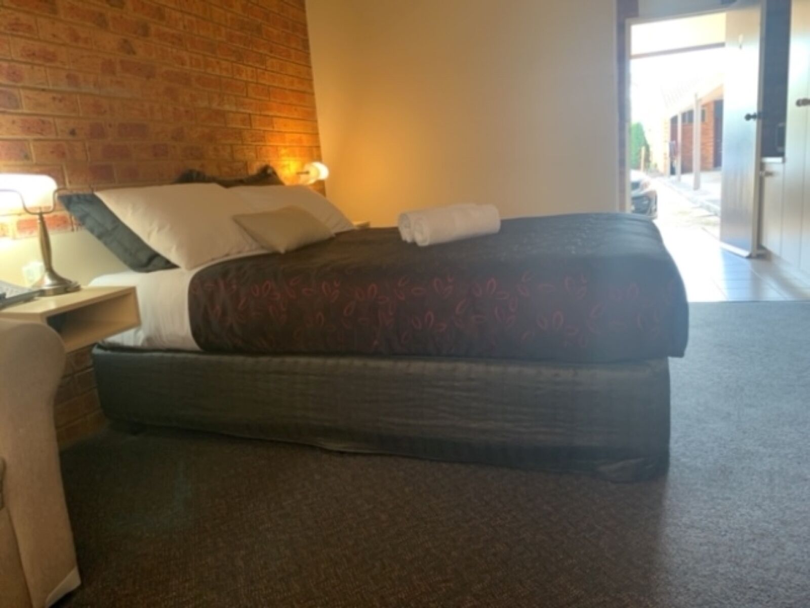 Habitación de hotel con pared de ladrillo, una cama con colcha estampada, una mesita de noche con lámpara y vista a la calle.