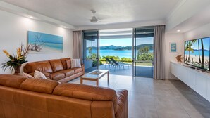 Un salon moderne avec un canapé en cuir, une table basse et un grand téléviseur à écran plat. La pièce est dotée de grandes portes vitrées qui donnent sur un balcon offrant une vue sur la mer et les montagnes.