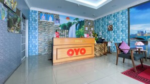 Ein Hotel-Rezeptionsbereich mit einem Holztisch mit der Aufschrift 'OYO', einer Rezeptionstheke, einem Stuhl mit violettem Sitzkissen und einem großen Fenster mit Blick auf die Stadt.