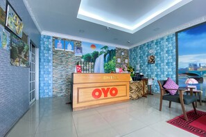 Un espace d’accueil d’hôtel avec un bureau en bois portant l’inscription « OYO », un comptoir de réception, une chaise avec un coussin violet et une grande fenêtre donnant sur le paysage urbain.