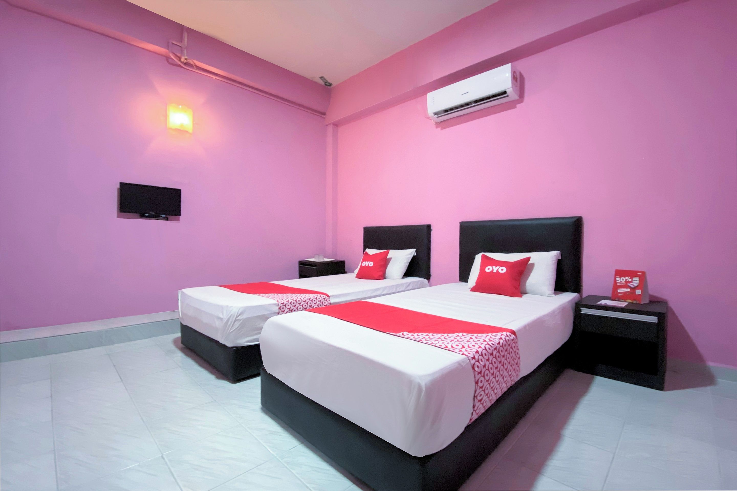 Habitación de hotel con dos camas, cada una con cabecero negro, ropa de cama blanca y detalles en rojo. Cuenta con un televisor de pantalla plana montado en la pared, un equipo de aire acondicionado y una pequeña mesa con un letrero rojo.
