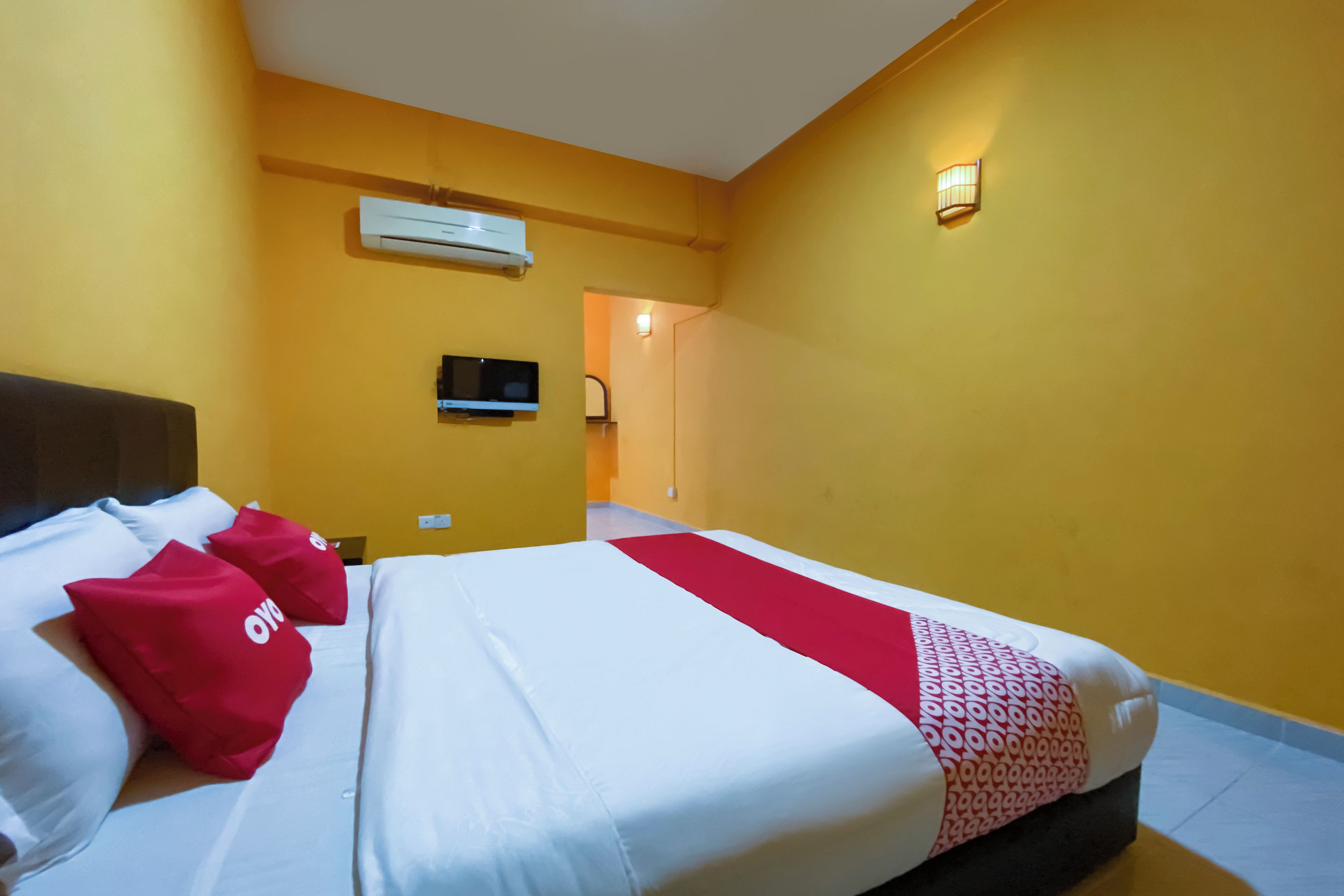 A hotel room with a bed, red pillows, a TV, an air conditioning unit, and a wall-mounted light fixture.