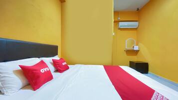 A hotel room with a bed, two red pillows with 'OYO' branding, a white and red bedspread, a black nightstand, and a wall-mounted air conditioning unit.
