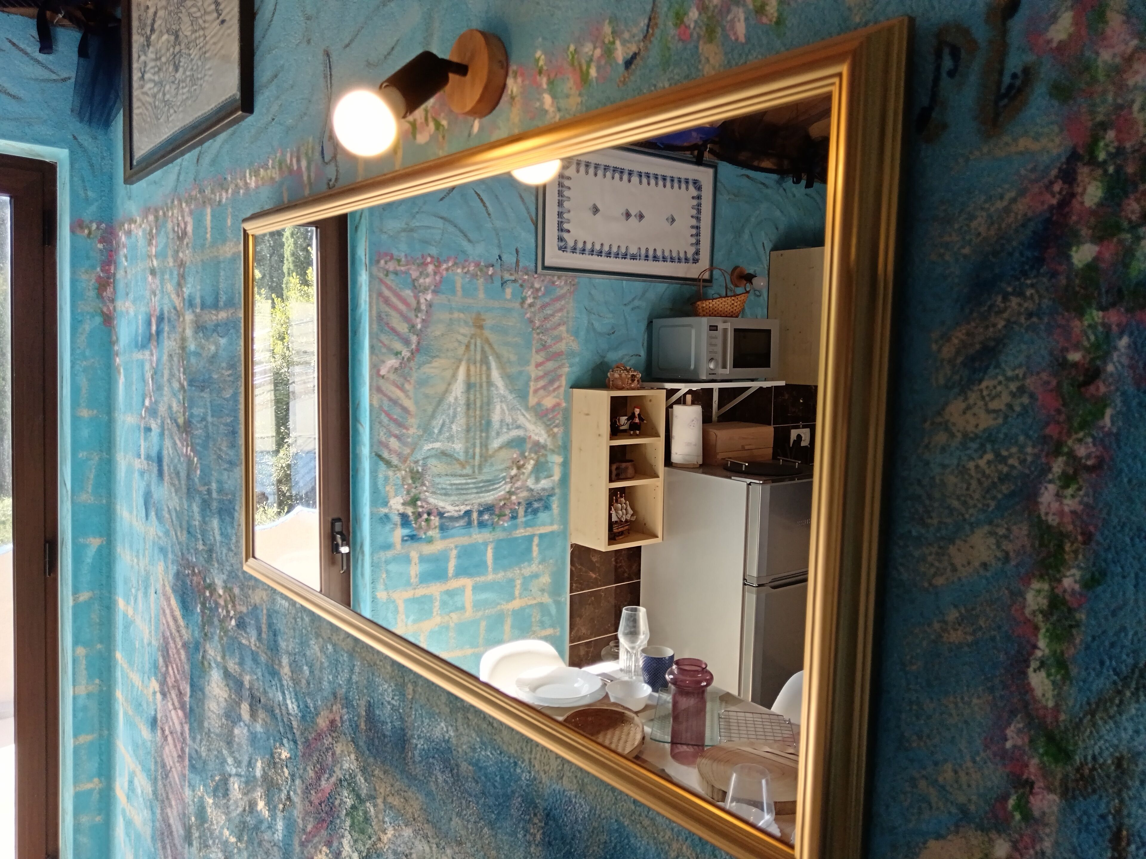 A room with a large mirror reflecting a kitchen area, a blue textured wall, and a framed artwork above the mirror.