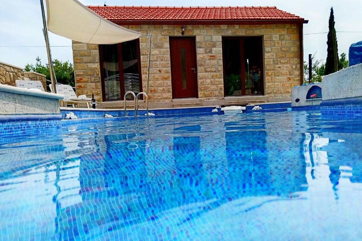 A swimming pool with clear blue water, a stone building with a red roof, and a white umbrella.