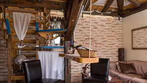 A rustic interior with a stone wall, wooden beams, a sofa, two leather chairs, and a hanging basket with a bottle and glasses.