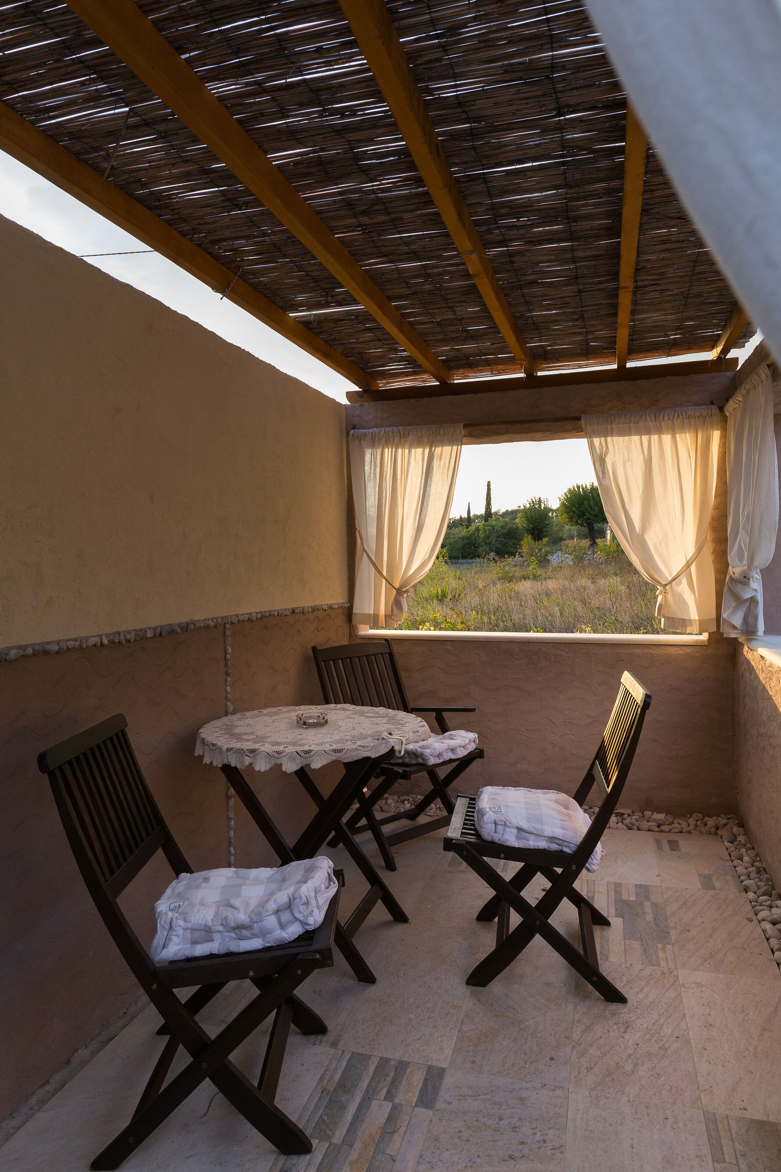 A covered patio area with a table and two chairs, a view of greenery outside, and a thatched roof overhead.