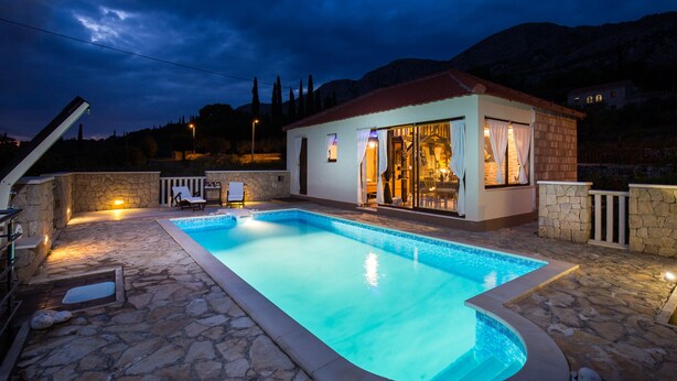 A swimming pool with a stone deck, a small building with a red roof, and a mountainous backdrop.