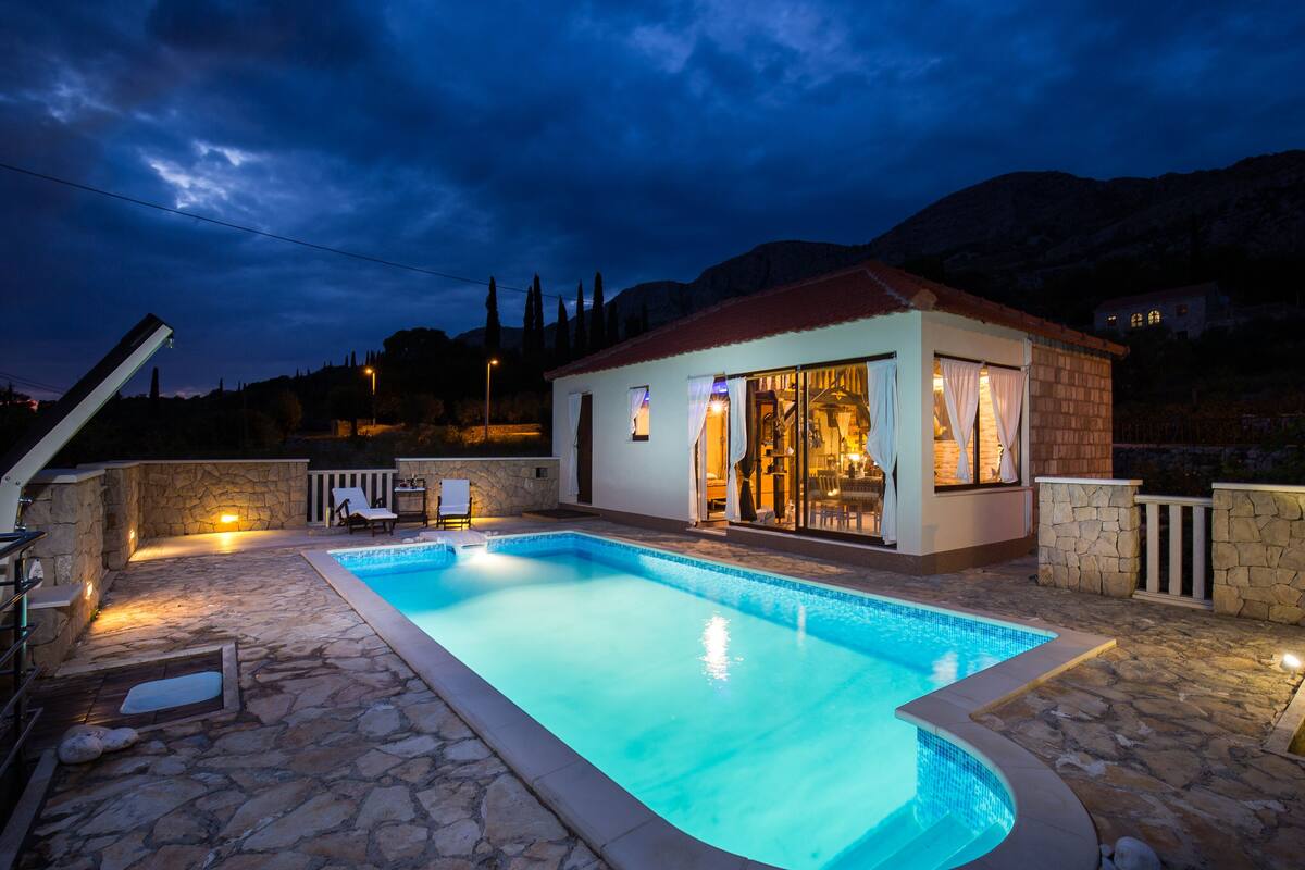 A swimming pool with a stone deck, a small building with a red roof, and a mountainous backdrop.