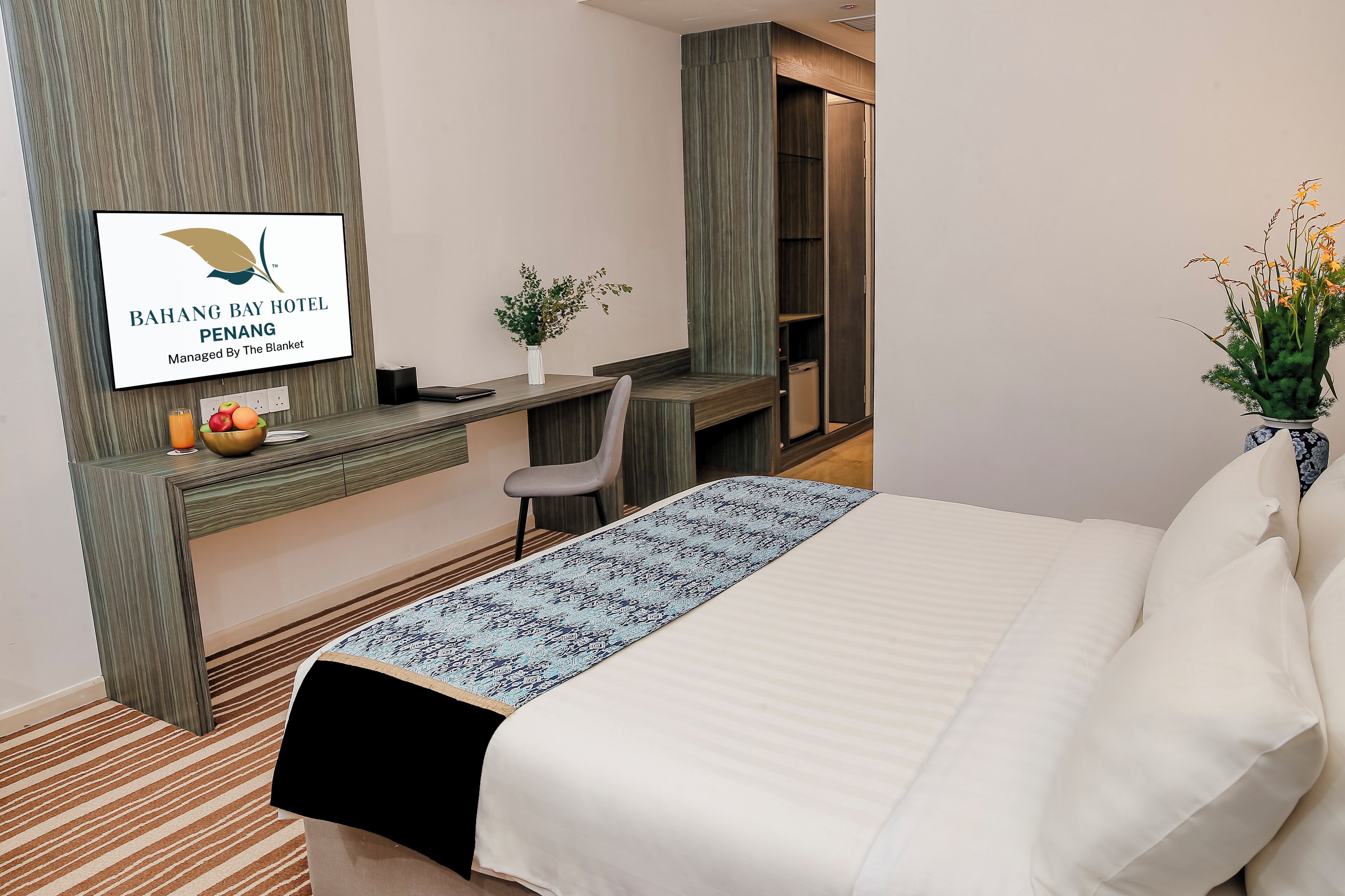 Hotellihuone, jossa on sänky, työpöytä, tuoli, hotellin nimen näyttävä televisio, maljakko kukkineen ja hedelmäkulho.