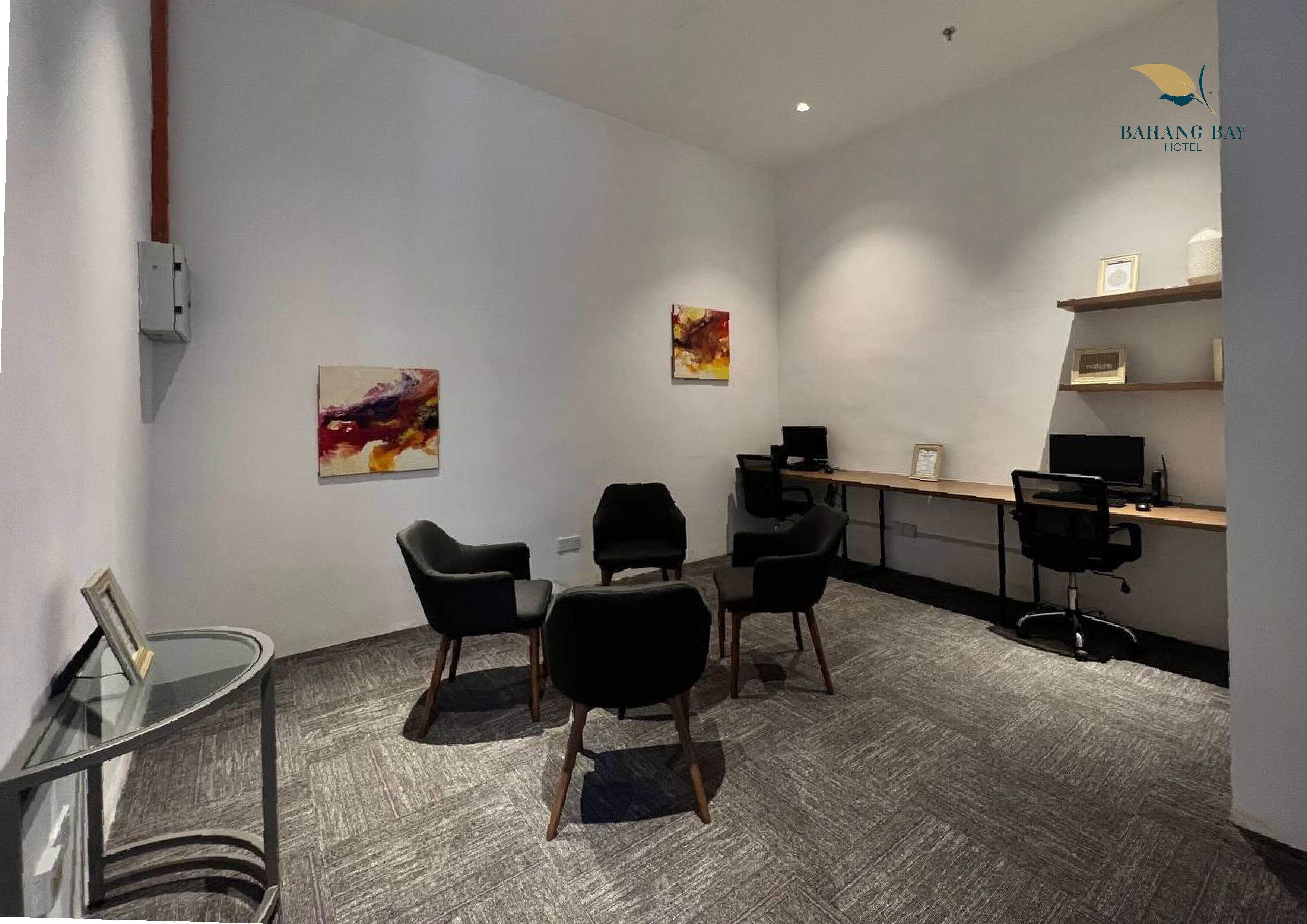 Una sala de reuniones moderna con una mesa de vidrio, cuatro sillas negras, dos cuadros, un escritorio con una computadora y una estantería de madera con un marco.