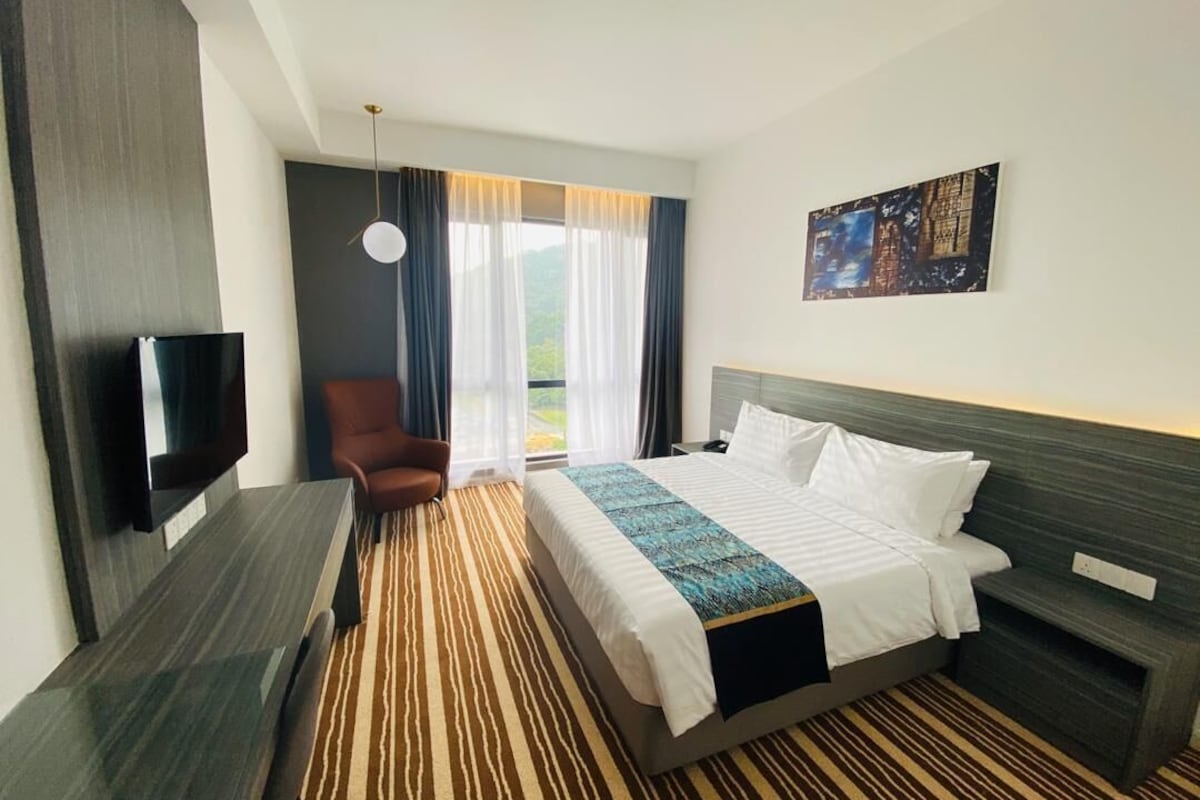 A hotel room with a bed, a TV, a chair, and a window with curtains.