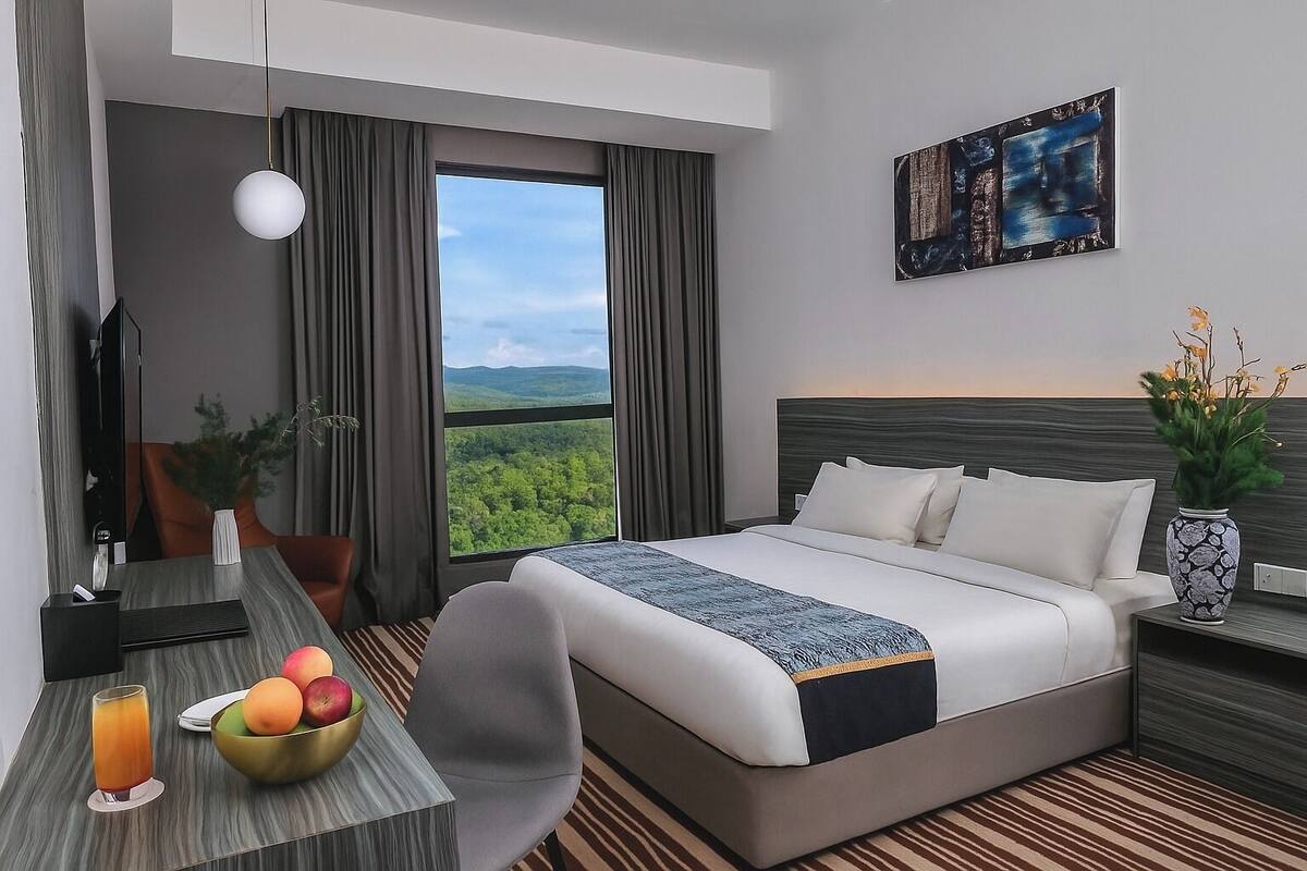 Une chambre d’hôtel avec un grand lit, un bureau agrémenté d’un panier de fruits, un vase de fleurs et un tableau au mur.