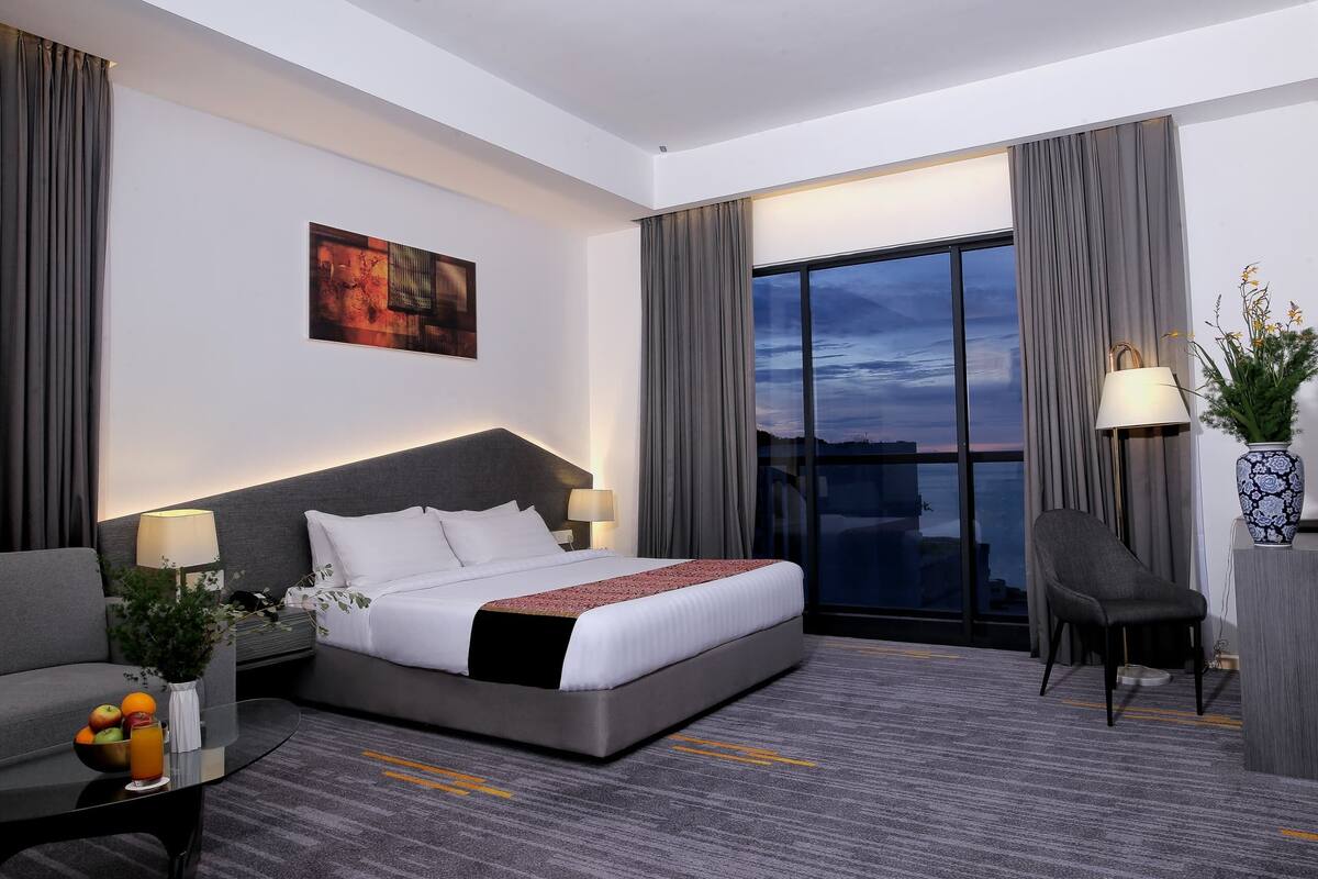 Une chambre d’hôtel avec un lit, un bureau, une chaise, une grande fenêtre avec des rideaux, un tableau au mur et un vase avec des fleurs.