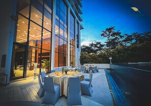 Un espace extérieur aménagé pour les repas, avec des tables et des chaises, une piscine et un bâtiment doté de grandes fenêtres.