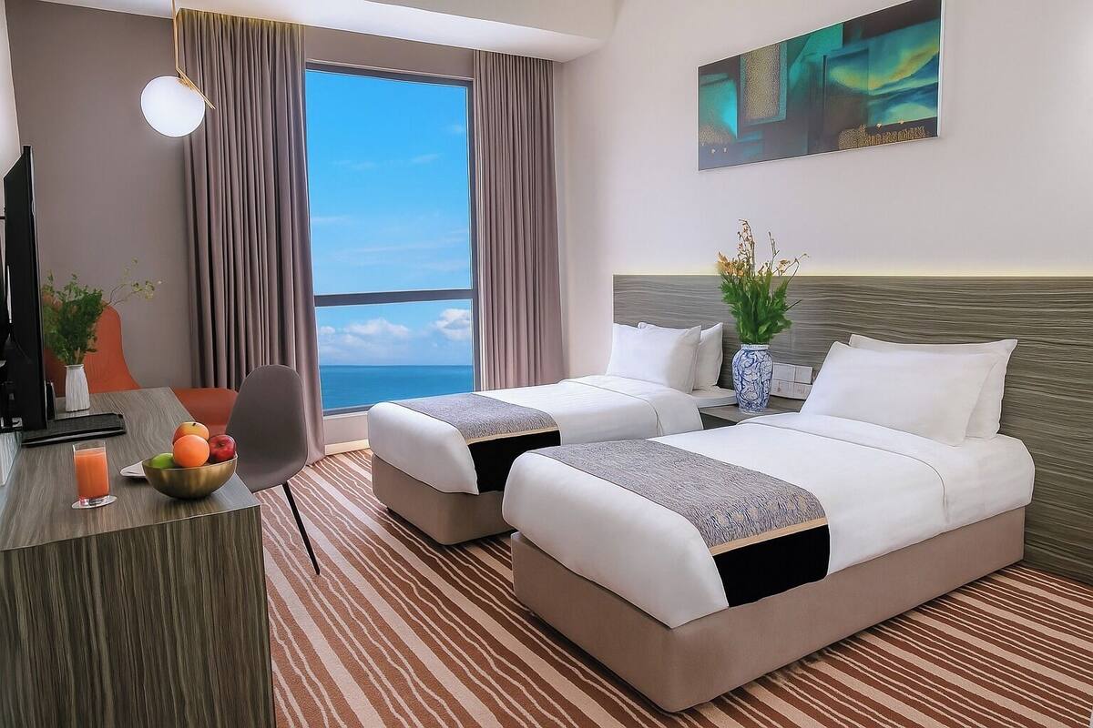 Une chambre d’hôtel avec deux lits, un bureau agrémenté d’un panier de fruits, un vase de fleurs et une grande fenêtre donnant sur l’océan.