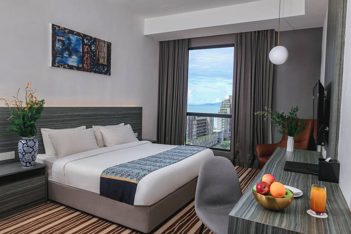 Une chambre d’hôtel avec un lit, un bureau agrémenté d’un panier de fruits et de boissons, un tableau au mur et une vue sur les bâtiments par la fenêtre.