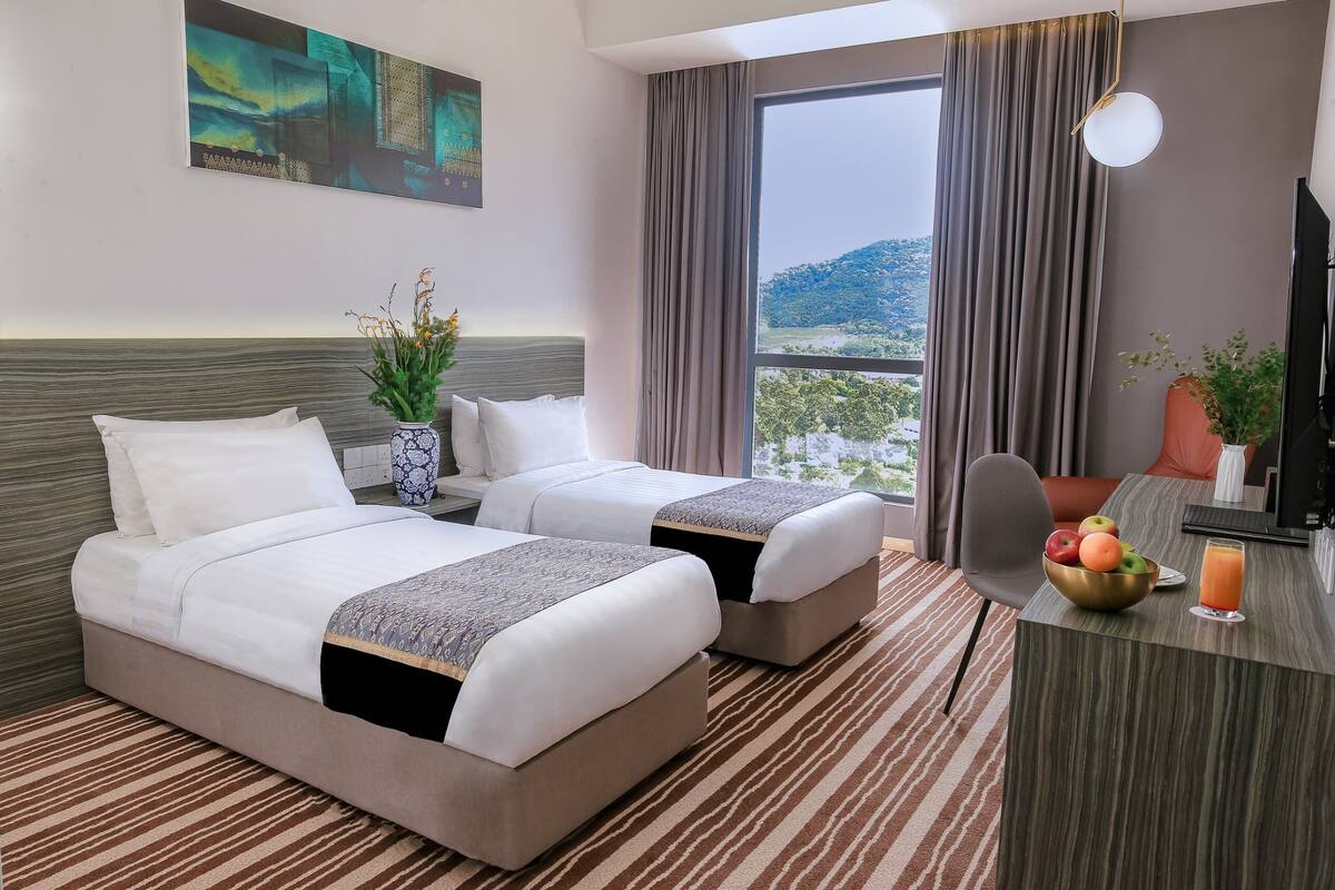 Une chambre d’hôtel avec deux lits, un bureau agrémenté d’un panier de fruits et d’une boisson, une grande fenêtre donnant sur un beau paysage et un tableau accroché au mur.