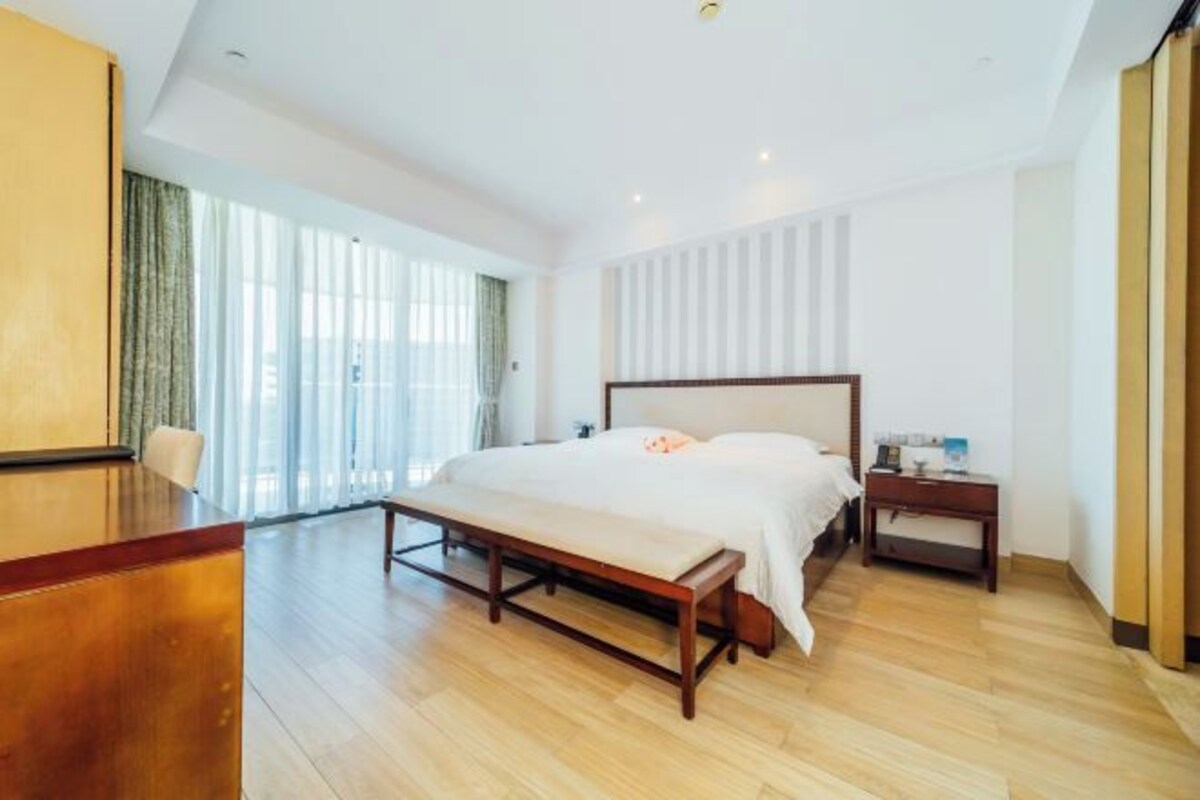 Ruime hotelkamer met een groot bed, houten vloer, een bureau en uitzicht op de stad door grote ramen.