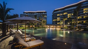 Un espace piscine aménagé dans un complexe hôtelier, avec des chaises longues et des parasols, entouré de grands immeubles dont les balcons sont illuminés.