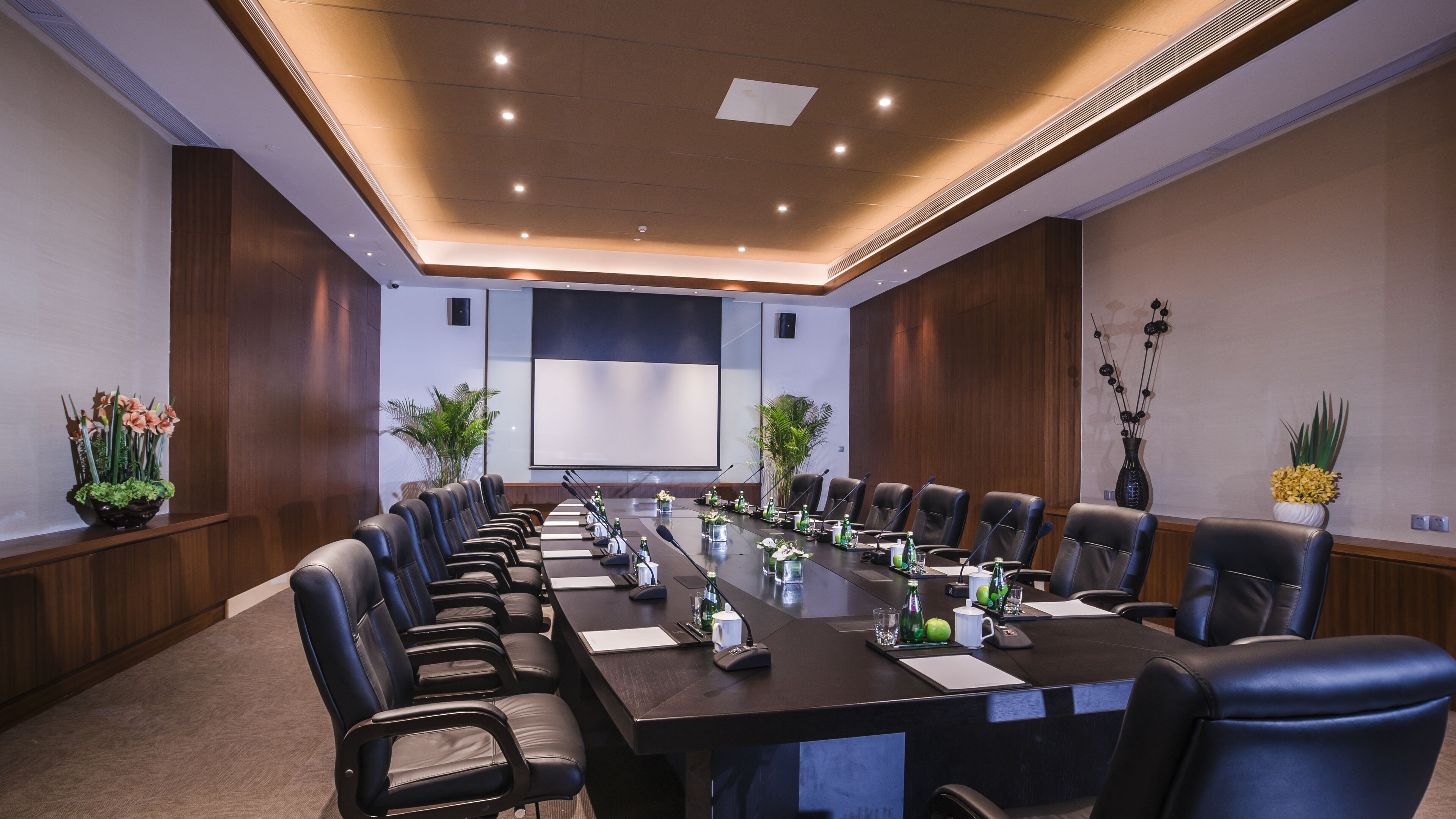 A conference room with a long table, black leather chairs, and a projector screen.