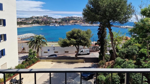 Vista costeira de uma varanda com vista para uma marina, com barcos, céu azul claro e um edifício com persianas azuis.