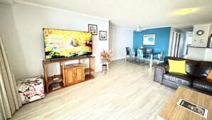 Ein Wohnzimmer mit Fernseher auf Holzständer, einer Essecke mit blauer Wand und einem Ledersofa mit gelbem Kissen.