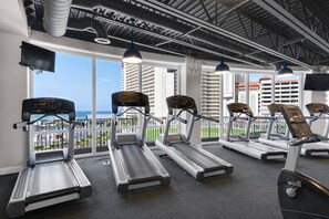 Uma academia com esteiras, bicicletas ergométricas e uma grande janela com vista para a paisagem urbana.