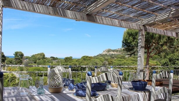 Un área de comedor al aire libre con vista al mar, cubierta por una pérgola a rayas y amueblada con manteles y sillas a rayas.