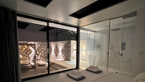 Un dormitorio moderno con una cama grande, una puerta de cristal que da a una zona de piscina, y una pared de piedra visible a través del cristal.