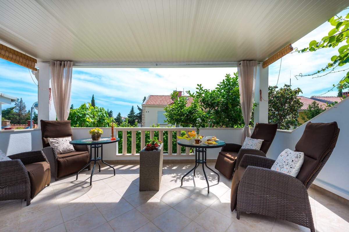 A covered outdoor seating area with wicker furniture, a small table with a vase, and a view of the neighborhood.