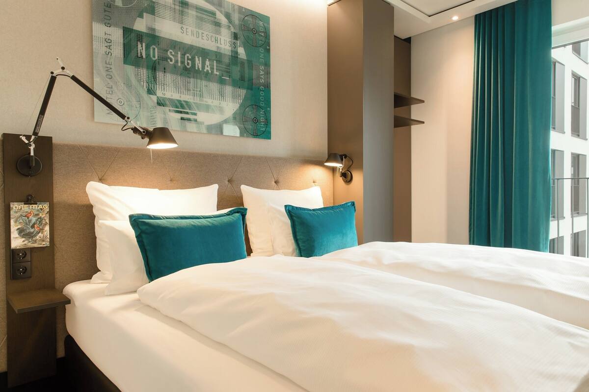 Habitación de hotel con una cama grande, dos lámparas de noche, un reloj de pared y una ventana con cortinas.