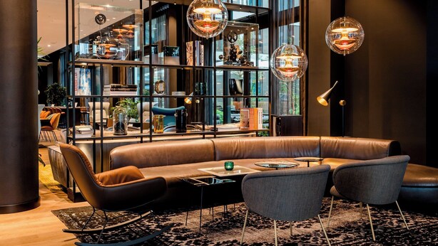 A modern lounge area with a leather sofa, armchairs, and a coffee table. There is a bookshelf filled with books and decorative items, and a staircase with glass railings leading to an upper level.