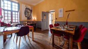 Un comedor con mesas de madera y sillas rojas, un ventanal con múltiples vidrios y un mural en la pared.