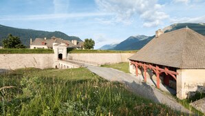 Un edificio histórico con un muro de piedra, un pequeño puente y montañas al fondo.