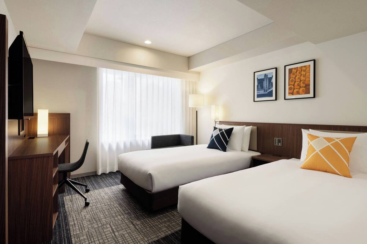 A hotel room with two beds, a desk, a chair, and a TV.