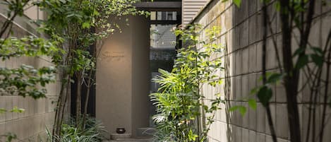 A narrow stone pathway flanked by greenery and a concrete wall, leading to a building entrance.
