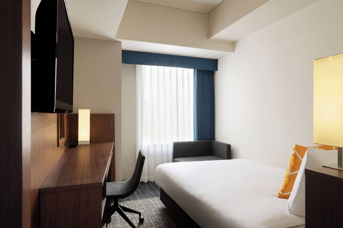A hotel room with a bed, a desk, a chair, a TV, and a window with curtains.