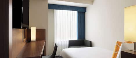 A hotel room with a bed, a desk, a chair, a TV, and a window with curtains.