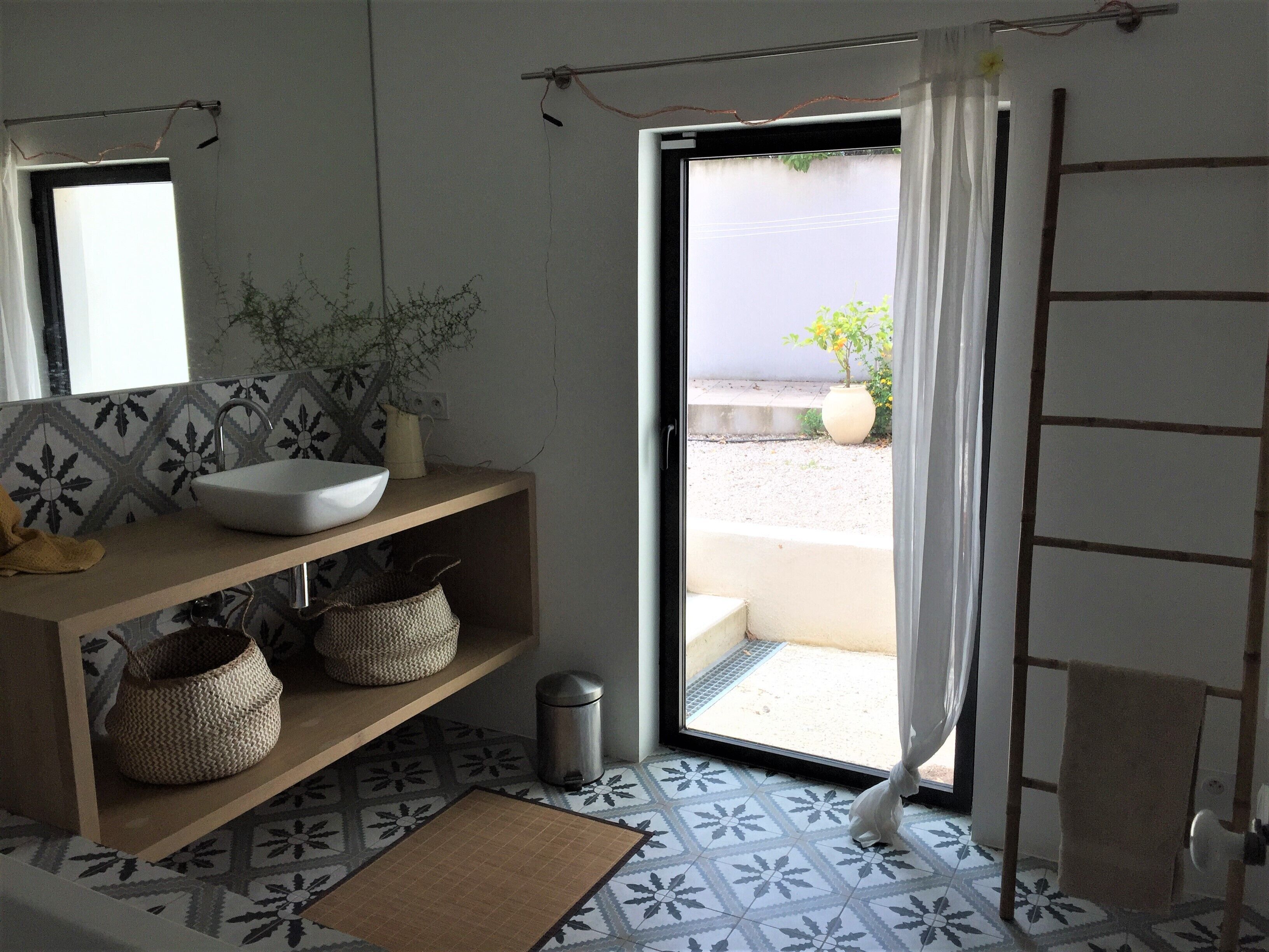 A bathroom with a white sink, wooden vanity, patterned floor tiles, a potted plant, and a view of the outside through an open door.